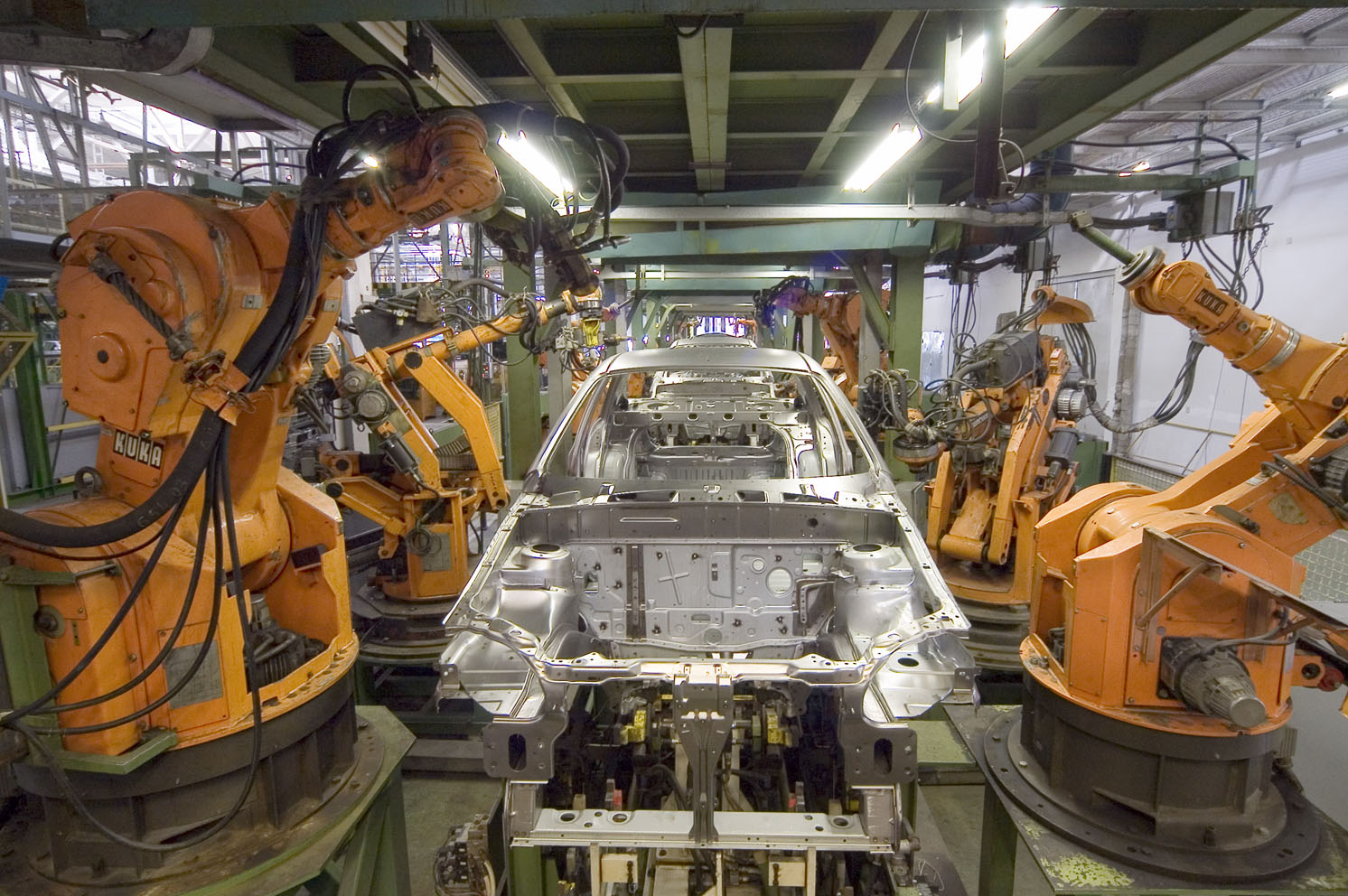 Orange KUKA industrial robot arms positioned around a silver car body in a factory.